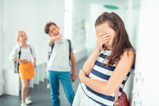 Two young school-aged children point and laugh at another student who is covering her face with her hands and is visibly upset.